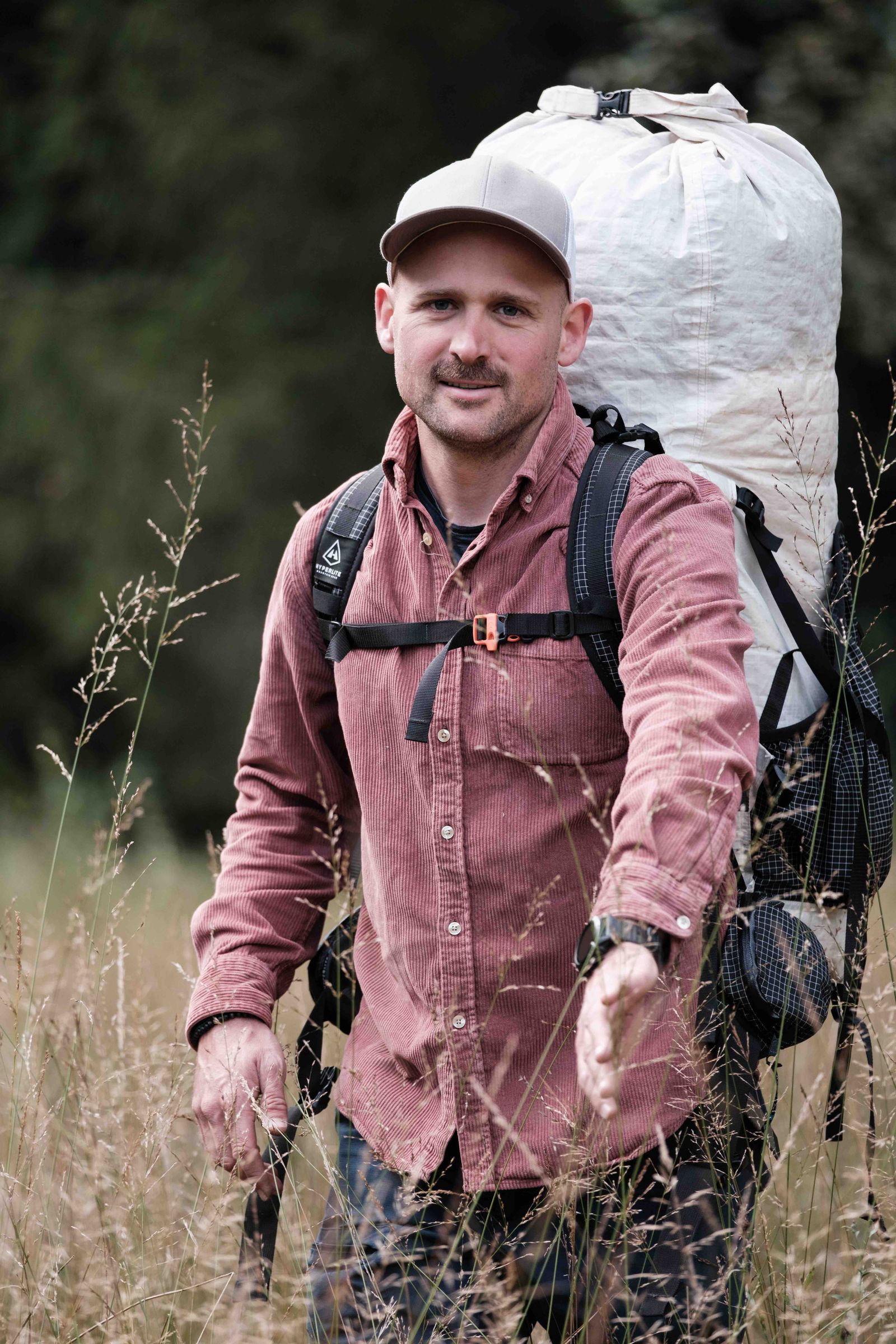 Sympathischer Outdoorguide Manuel Seiler mit grossem weissen Rucksack, Basecap und rotem Manchesterhemd mitten im Feld